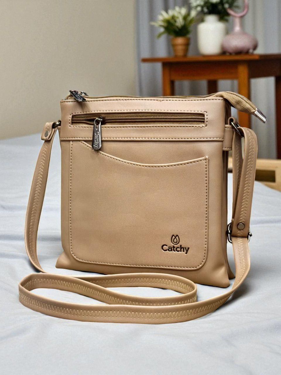 Beige Catchy multi-compartment crossbody bag on a textured bedspread in a modern bedroom.