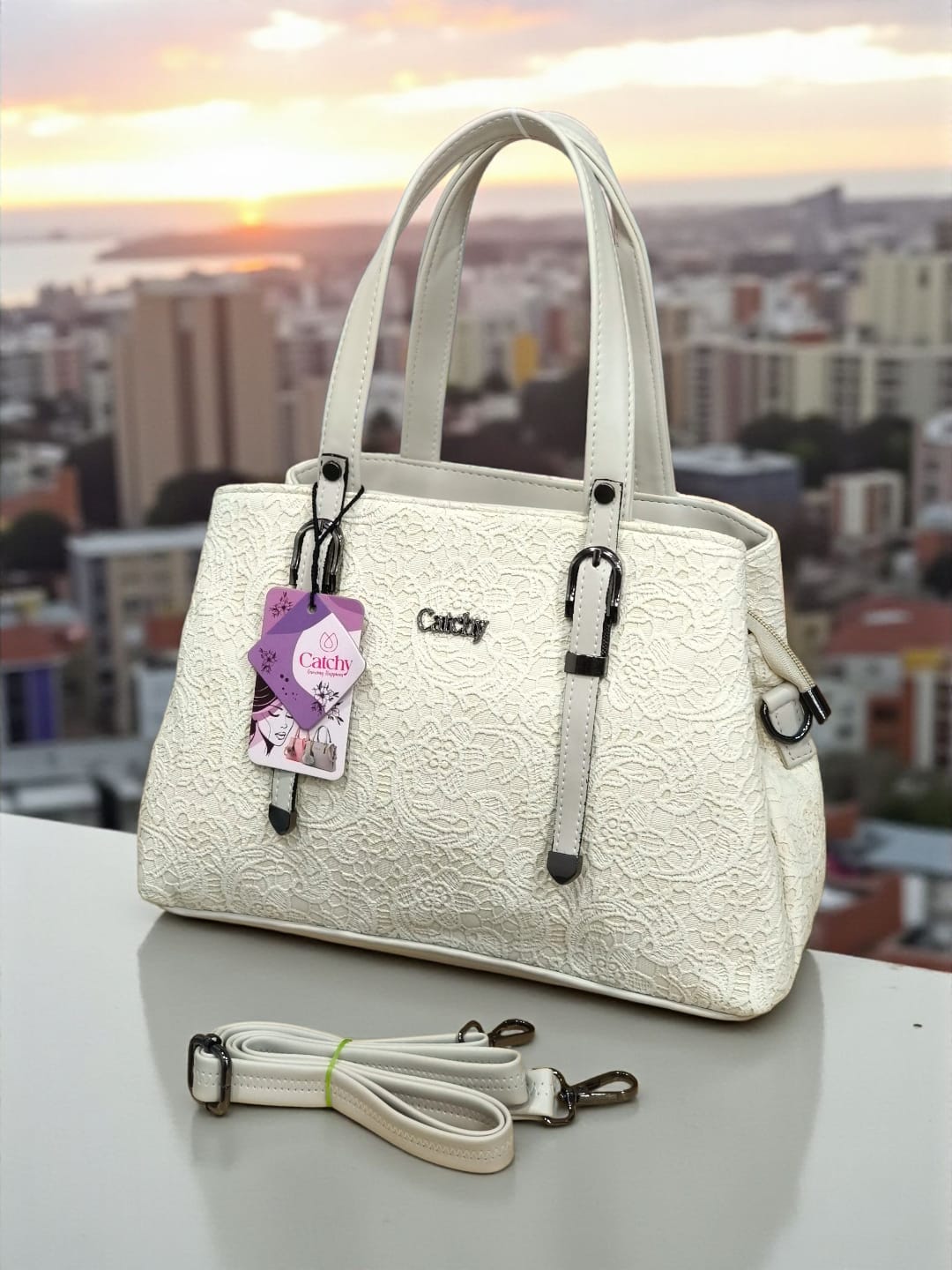 Stylish white floral embossed TRENDBUY handbag with detachable sling strap, displayed on a railing with a city skyline at sunset.