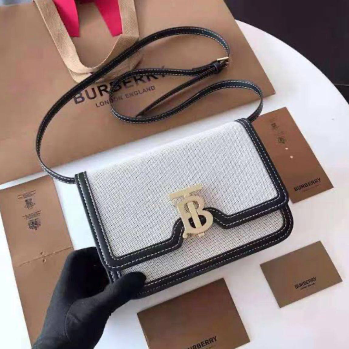 Stylish white canvas Burberry style crossbody sling bag with sleek black leather trim and gold TB logo, presented with branded accessories and packaging.
