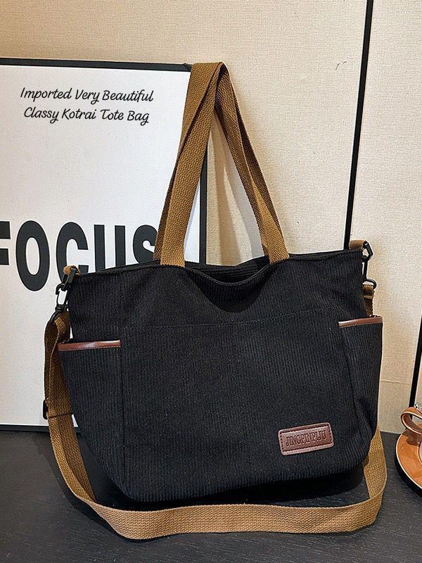 Front view of a black corduroy tote bag with brown straps, side pockets, and a leather brand patch, highlighting its imported, beautiful, and classy design.