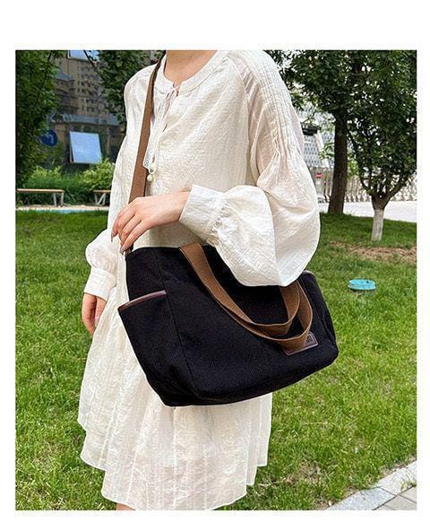 Woman in a light-colored dress carrying a black corduroy tote bag on her shoulder, walking outdoors, demonstrating its comfortable shoulder fit.