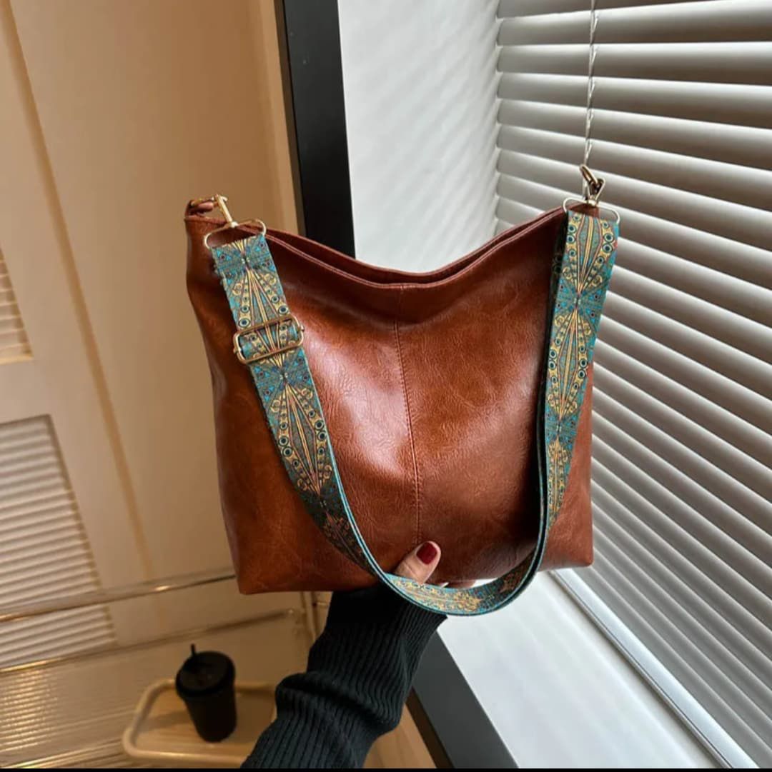 Hand holding a brown TRENDBUY PU leather tote bag with a colorful patterned strap, by a window with blinds.
