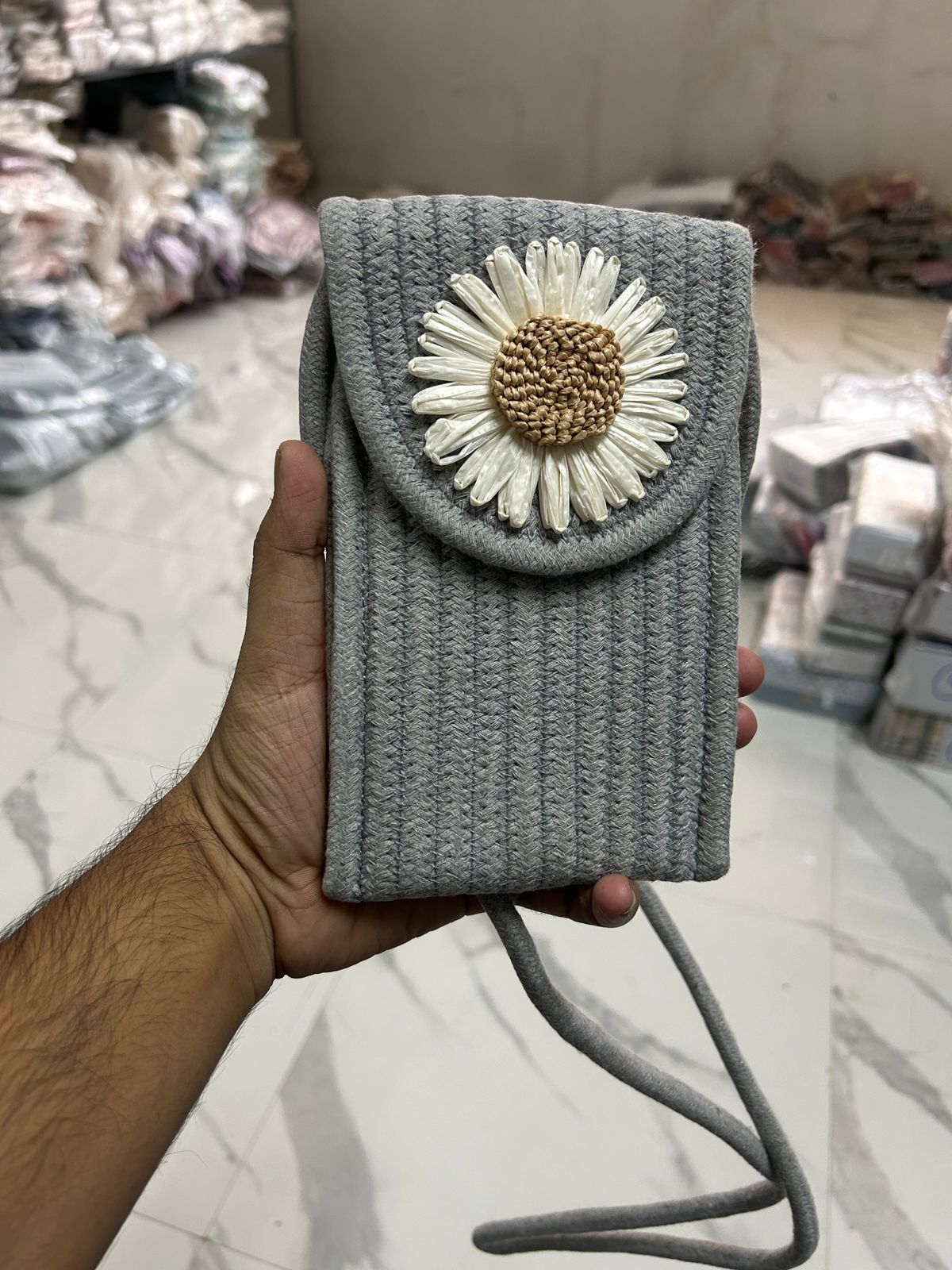 Grey braided crossbody phone bag with a decorative white flower embellishment and a gold center, held in hand.