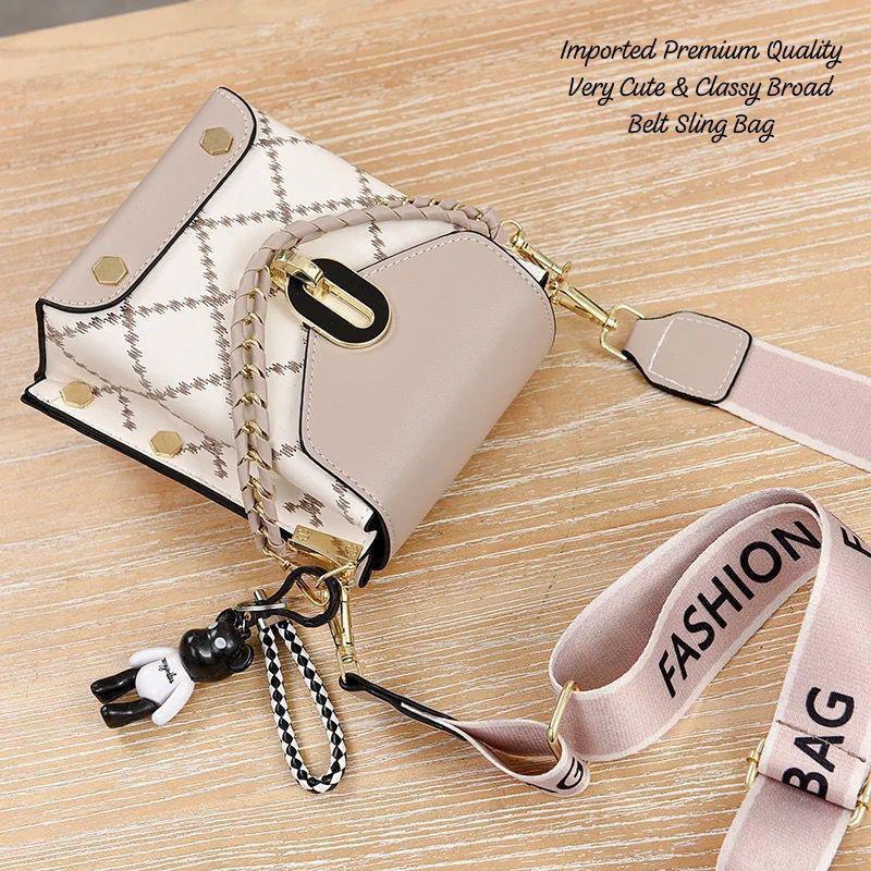 TRENDBUY beige and white broad belt sling bag with a decorative chain and a black teddy bear keychain, featuring a 'FASHION BAG' strap on a wooden surface.