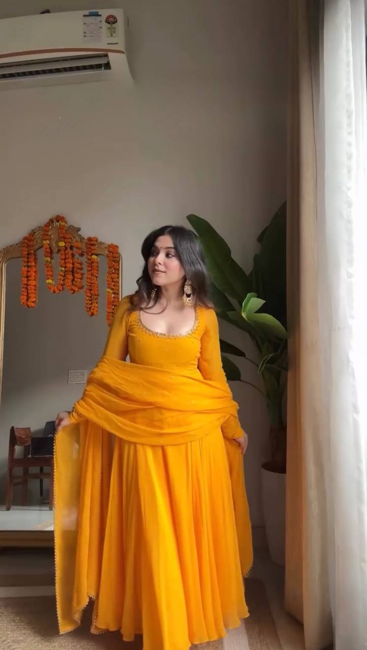 Woman in a vibrant yellow Faux Georgette Anarkali suit, posing gracefully near a window.