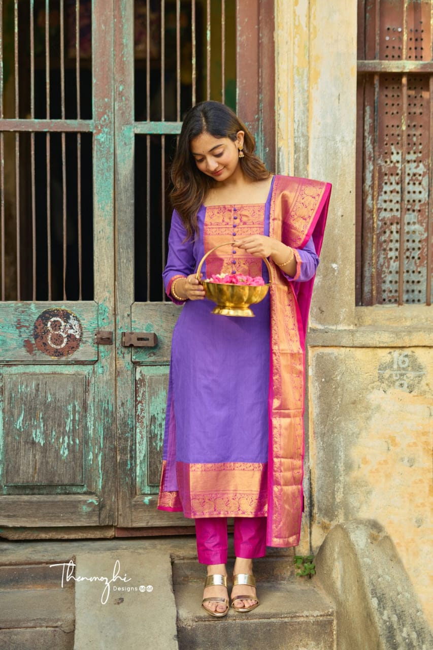 Woman in purple Banarasi silk suit set with pink and gold dupatta, holding a flower basket, standing by a rustic door.