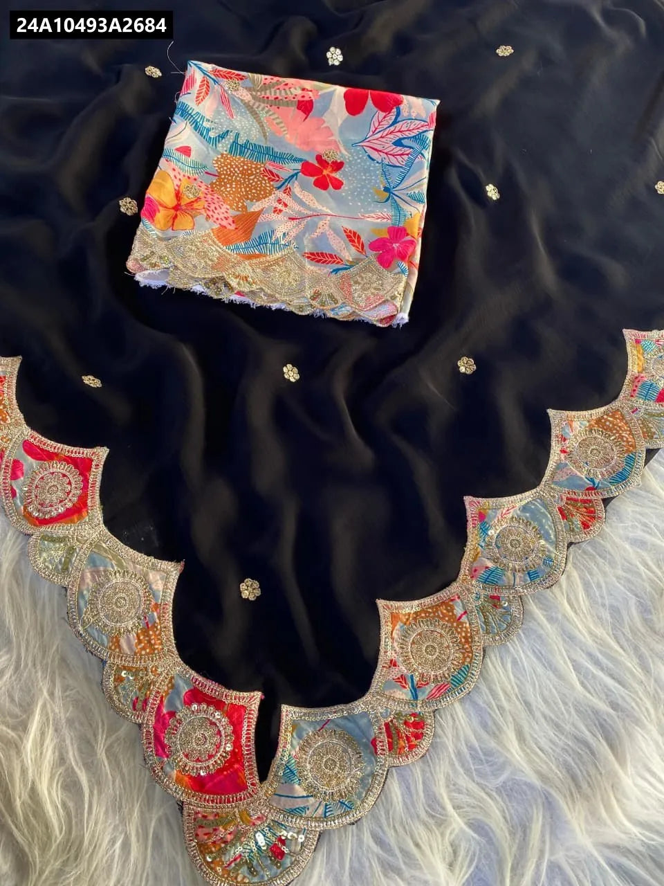 Top-down view of black satin georgette saree with appliqué cutwork border, delicate sequence buttis, and folded floral print contrast blouse on a white furry surface.