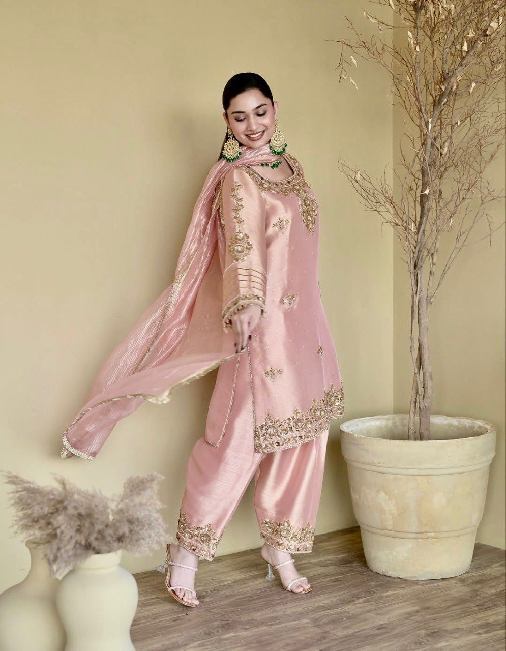 Woman posing gracefully in a blush pink natural silk plazzo suit set with intricate gold embroidery and pearl work, perfect for festive occasions.