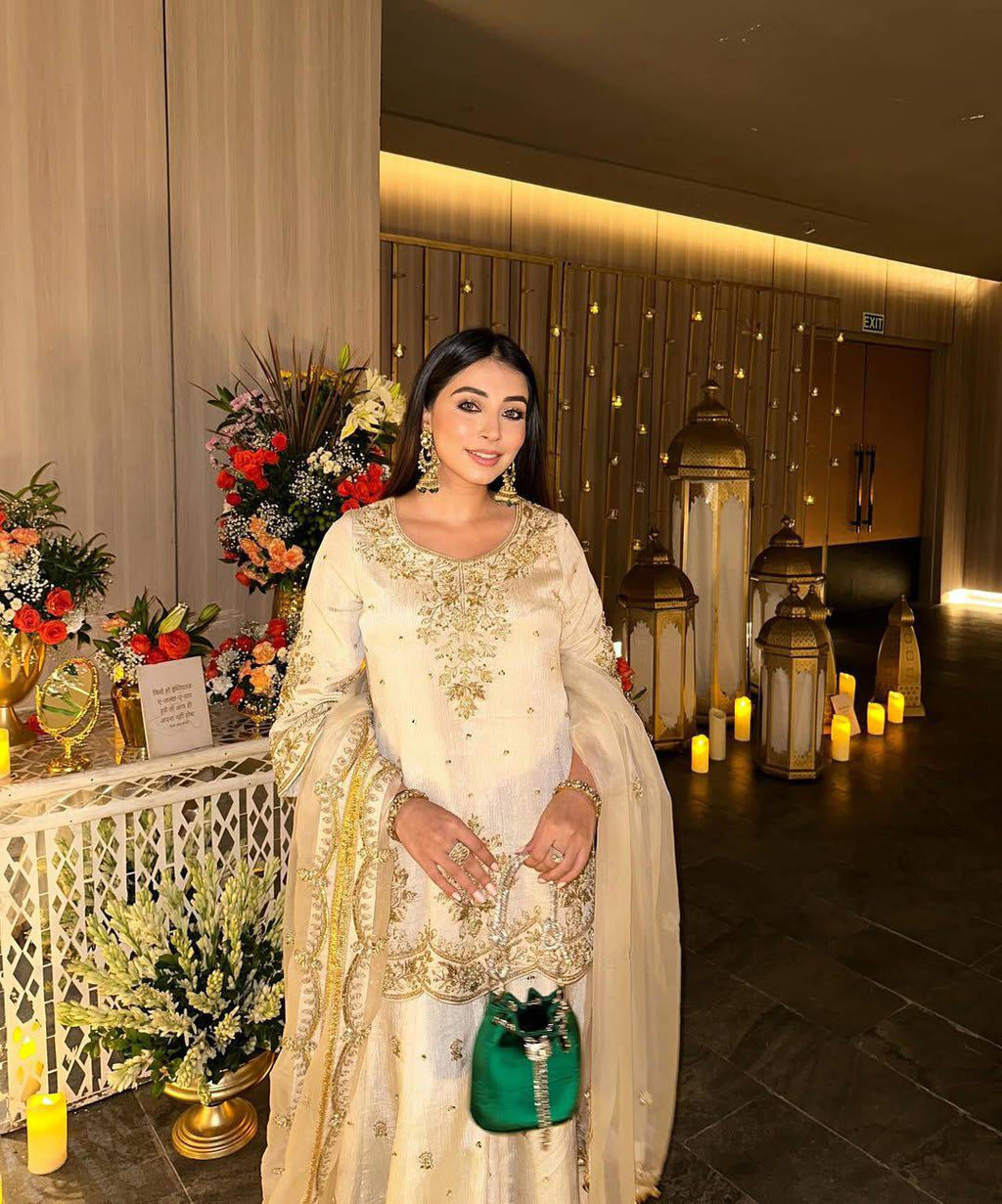 Full-length shot of a woman smiling, wearing an elegant cream Chinnon Silk Kurti Plazzo Set with intricate gold sequin and zari embroidery, holding a green clutch, amidst festive decor.