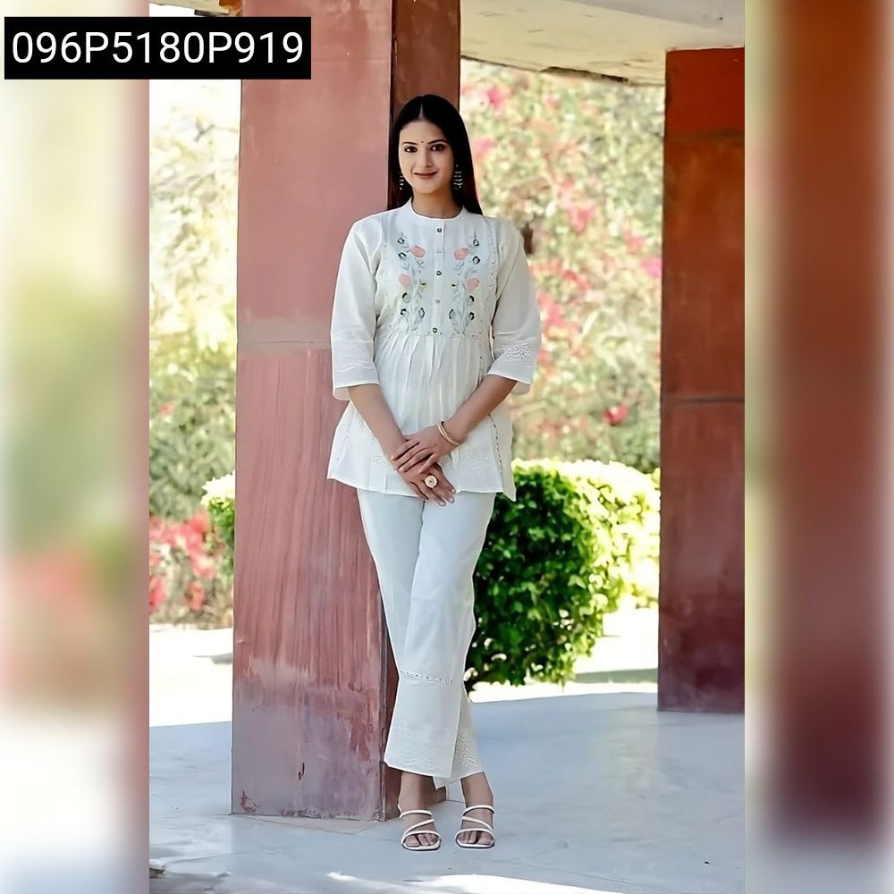 Full shot of a woman wearing a white cotton dobby shiffli embroidered tunic and matching pants, smiling gently against an outdoor backdrop.