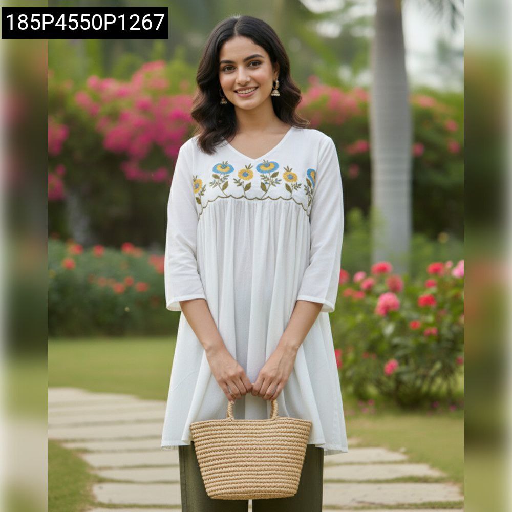 Women's white cotton embroidered tunic top with olive pants and straw bag, smiling at camera in a garden setting.