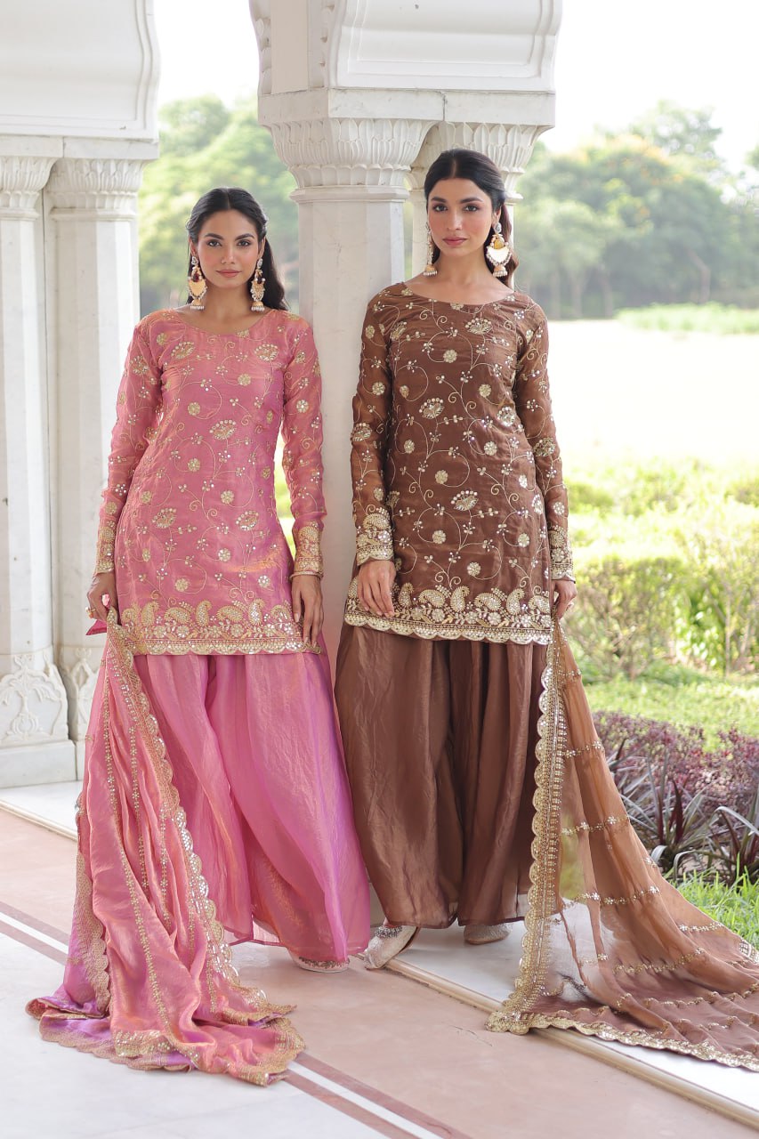 Two women gracefully posing in designer readymade sharara sets, one in pink and one in coffee brown, featuring intricate embroidery.