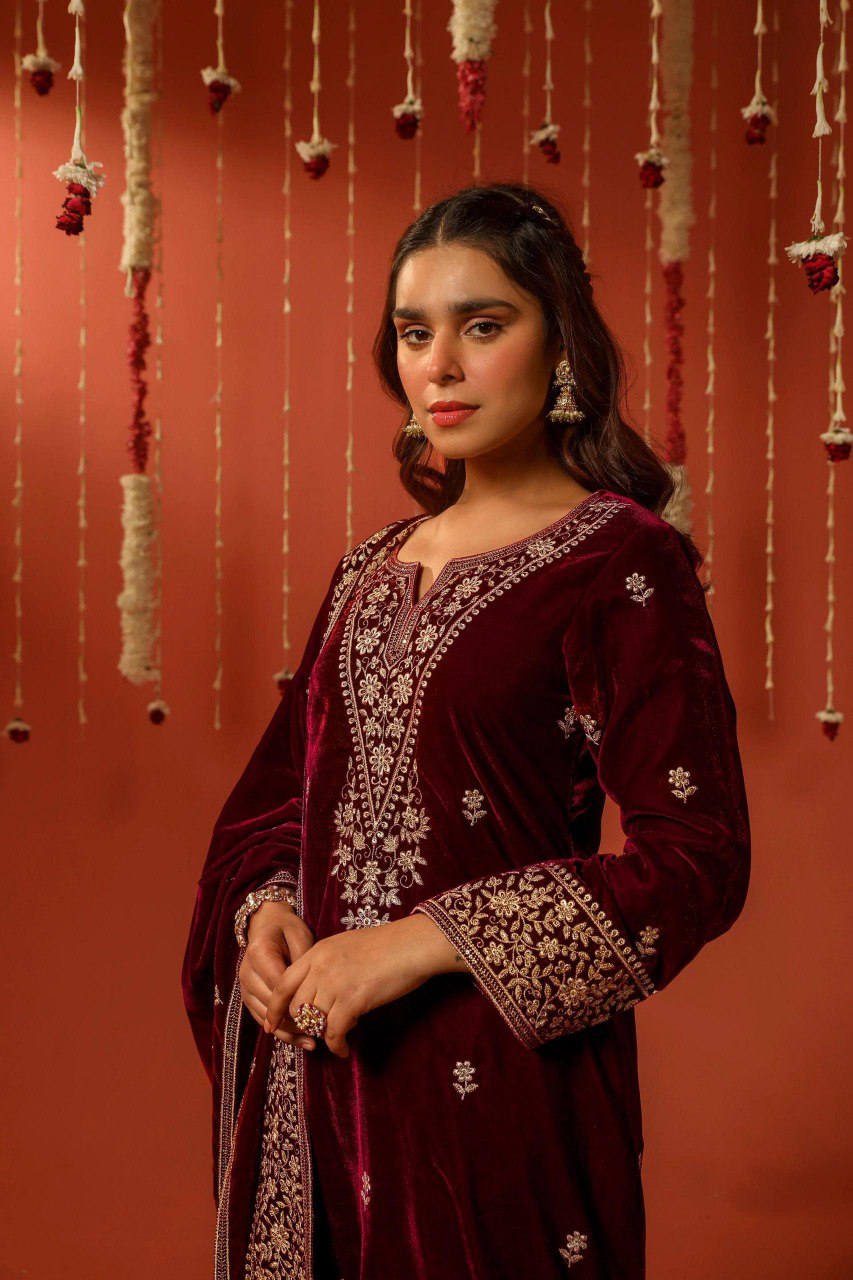 Woman in maroon designer velvet suit with intricate zari and sequin embroidery.