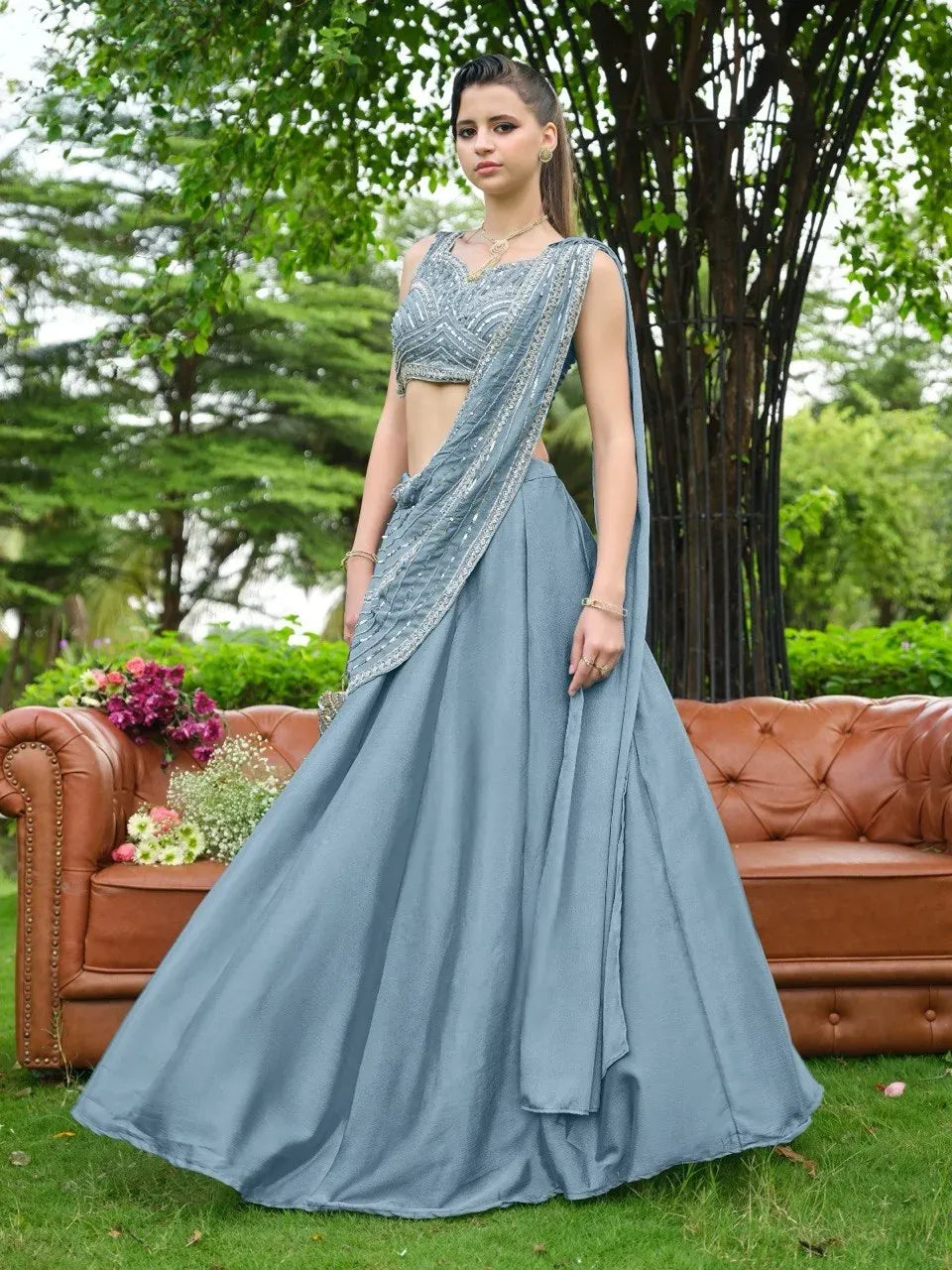 Woman in a dusty blue chinnon lehenga standing outdoors on green grass near a leather sofa