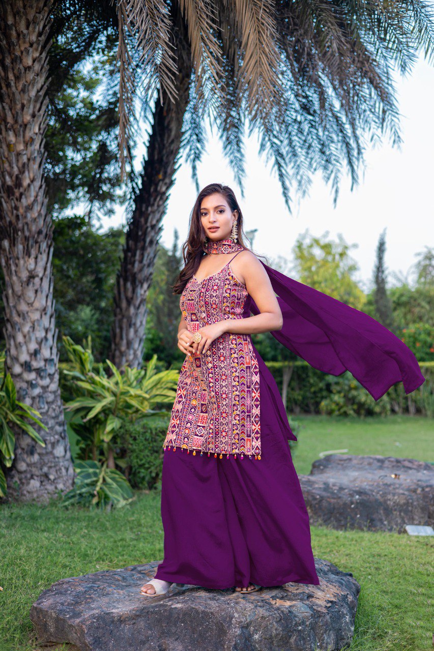 Woman in a vibrant purple embroidered Chinon Sharara suit with matching dupatta by TRENDBUY, posing outdoors amidst lush greenery.