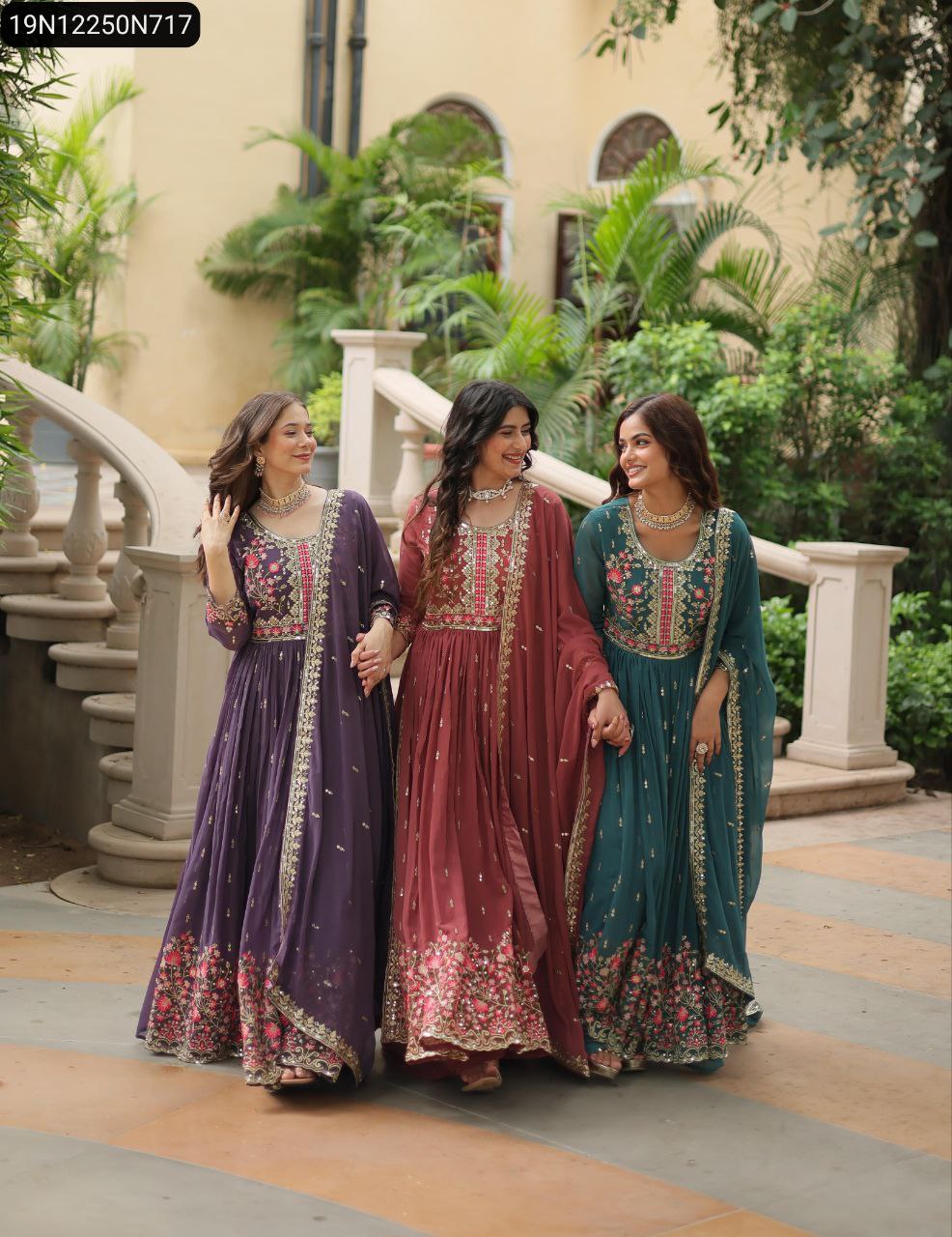 Three women in purple, wine red, and teal embroidered Faux Georgette Sharara Suits, walking hand-in-hand in a garden setting.