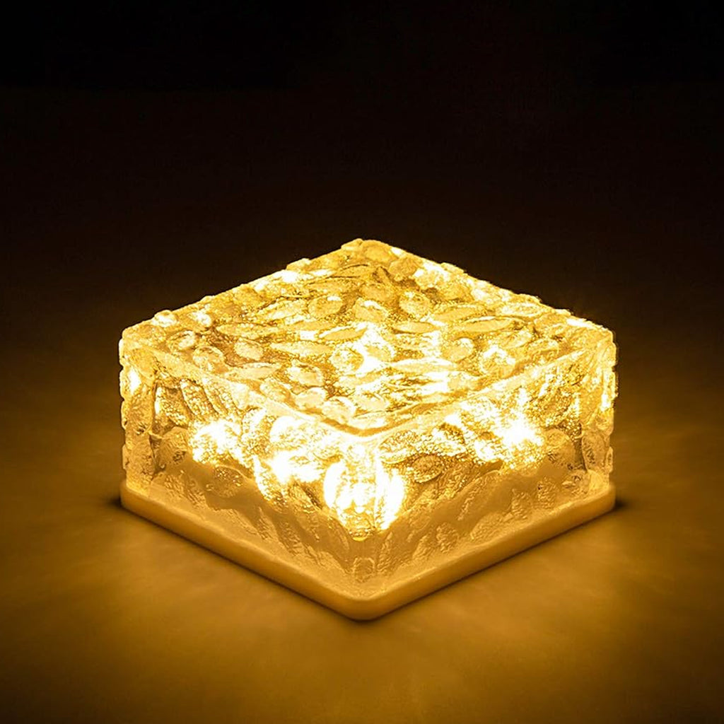 Close-up of a square frosted glass solar brick emitting a warm yellow light on a dark stone pathway