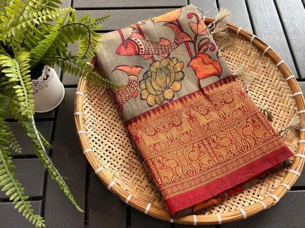 Folded grey linen saree with kalamkari fish print on a woven tray next to a potted fern