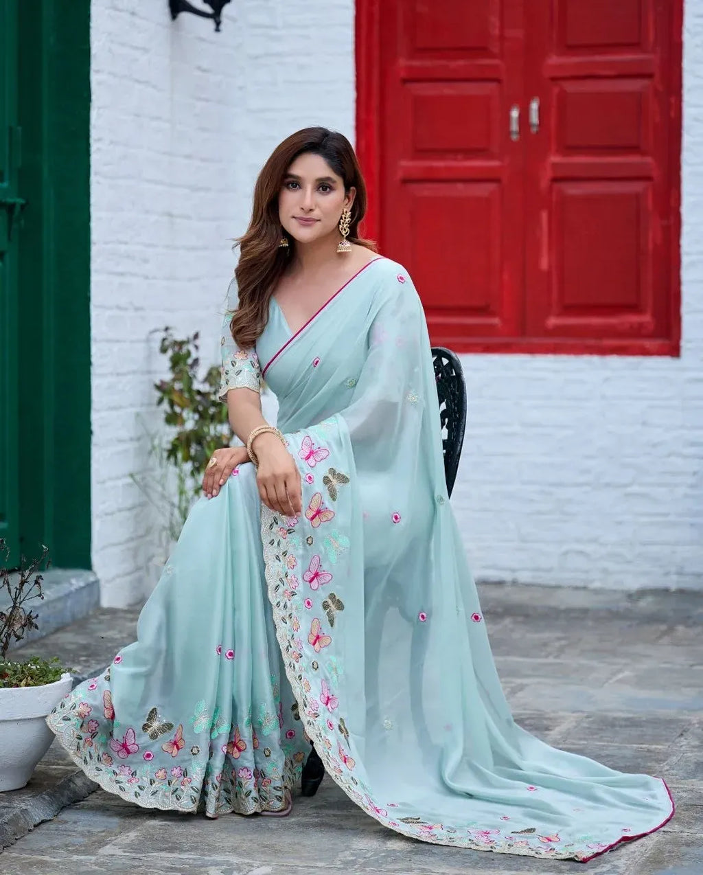 Light blue rangoli saree with colorful butterfly embroidery on model seated against white brick wall