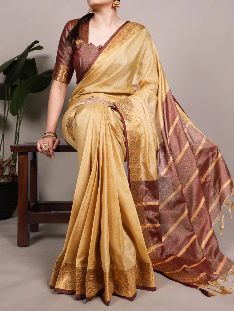 Model wearing gold poly cotton saree with brown zari border sitting on a wooden bench