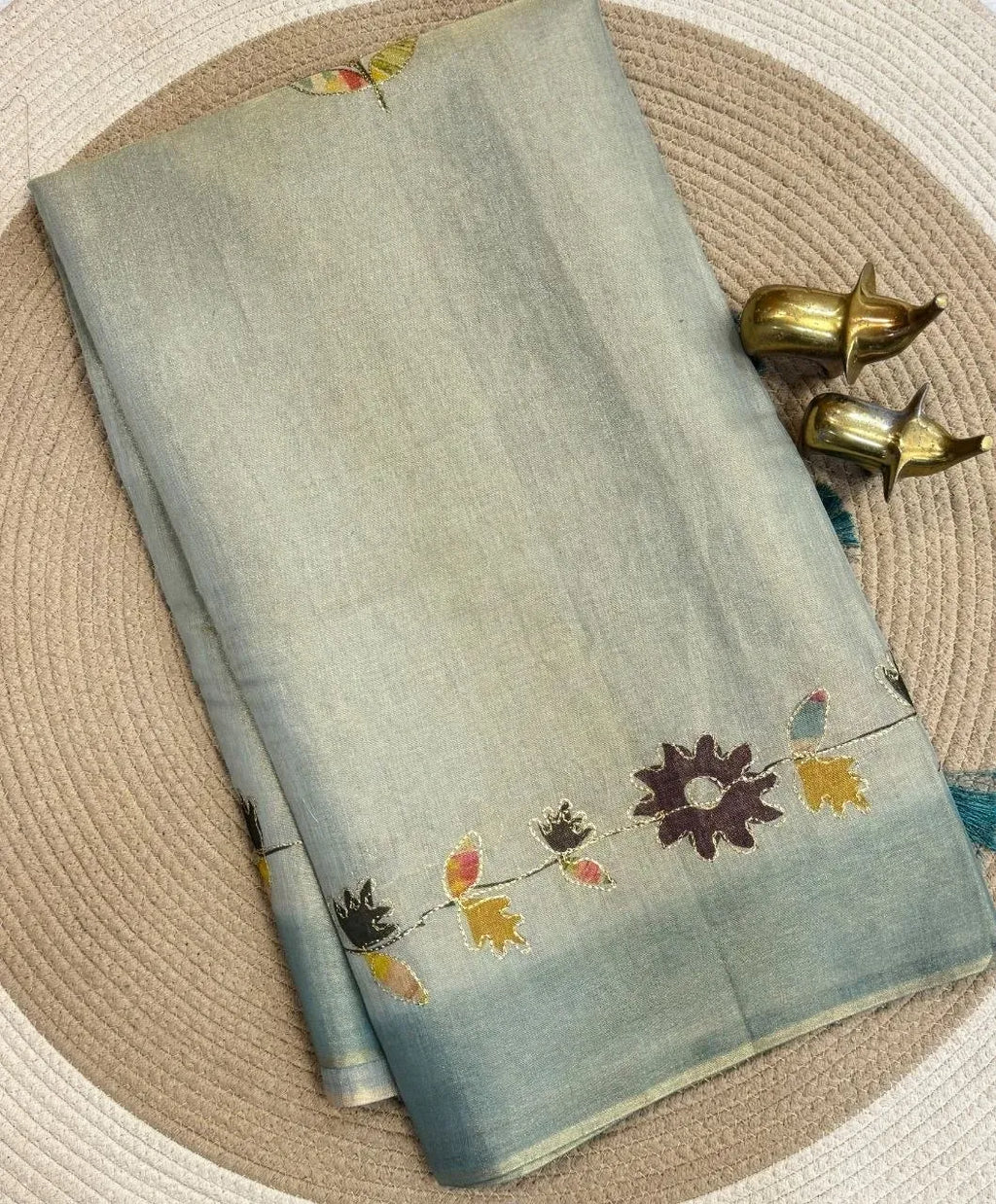 Folded soft grey tissue saree with floral zari embroidery and brass elephant figurines on a woven mat