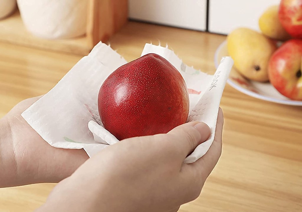 Person holding a red apple wrapped in paper towel on a wooden surface.