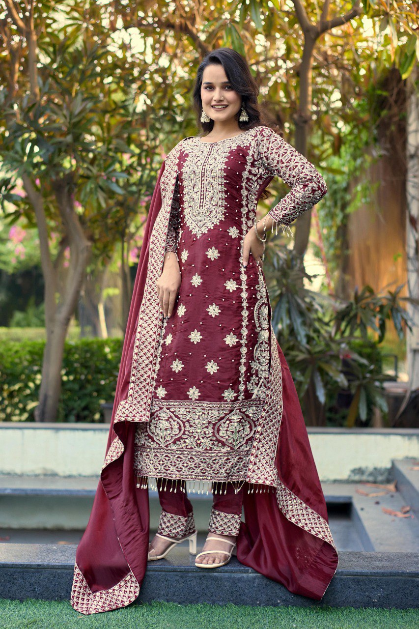 A woman smiling, wearing a maroon GMY embroidered suit set with sequence work and matching dupatta, posing elegantly outdoors.