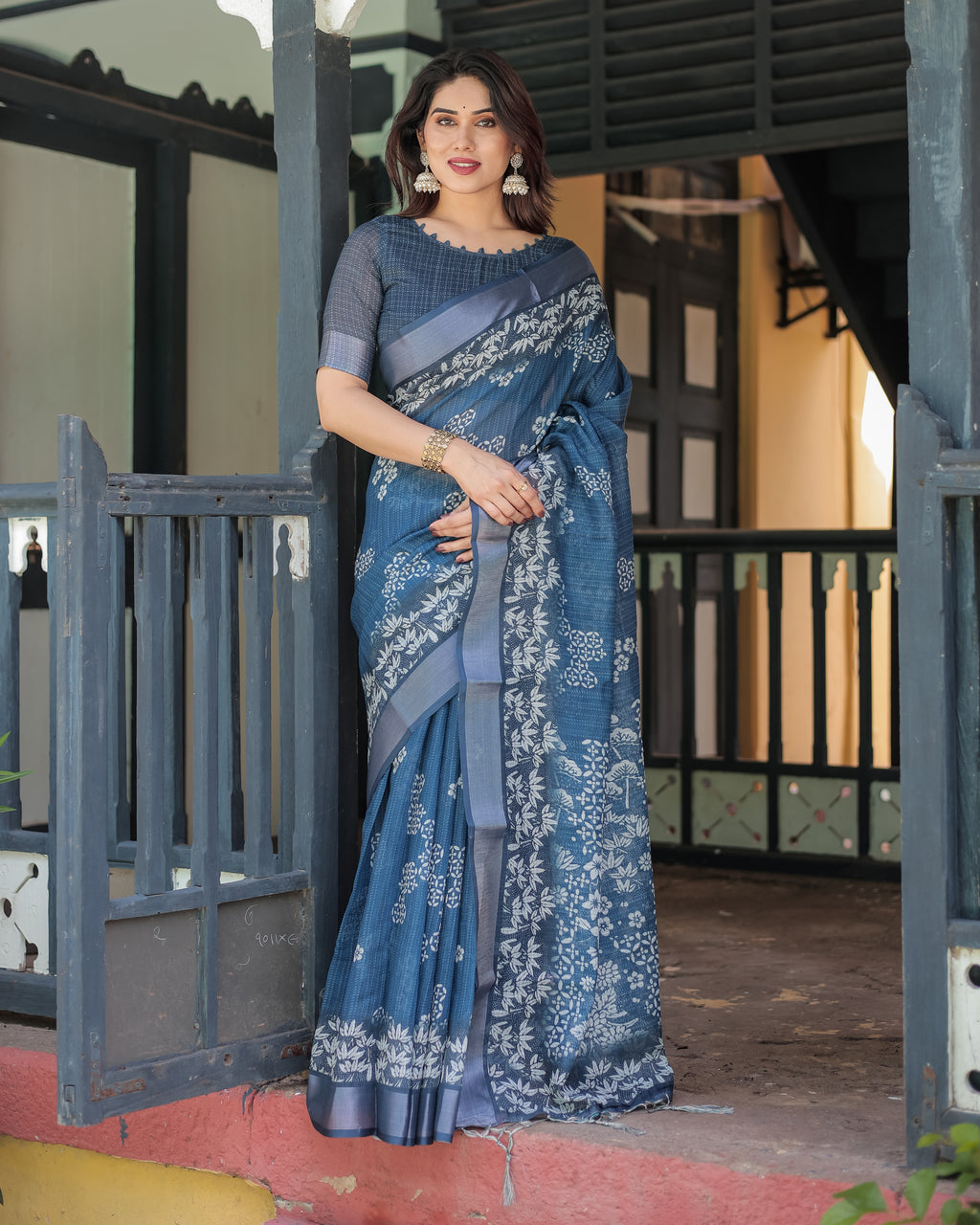 Full body shot of a woman modeling a deep blue floral linen saree with an elegant blouse, standing gracefully at an entrance.