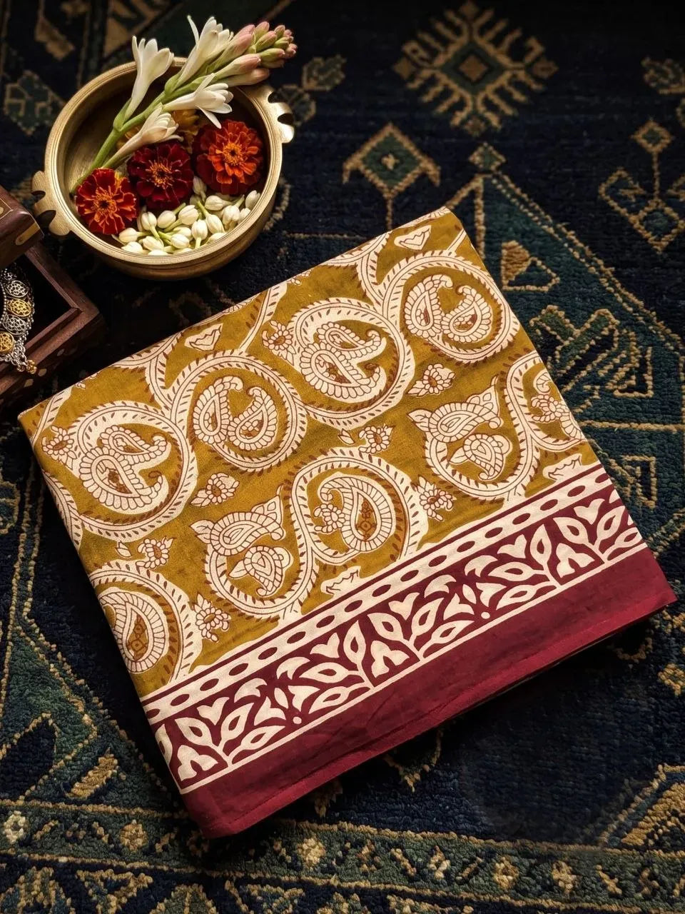Folded mustard yellow artisan cotton saree with floral print next to brass bowl of flowers