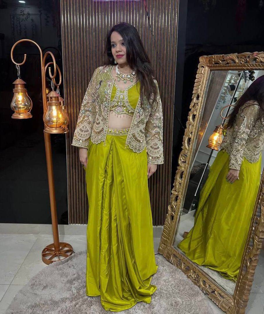 Full body shot of a woman modeling a lime green Indo-Western outfit with an embroidered sheer koti, standing beside a decorative lamp.
