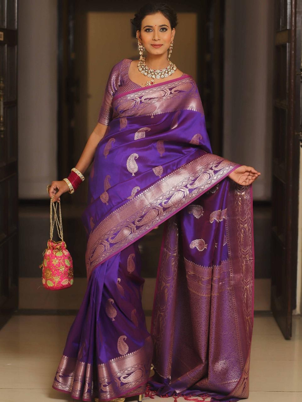 Woman wearing a vibrant Purple Lichi Silk Gold Zari Saree with intricate jacquard work and a matching blouse, holding a potli bag.