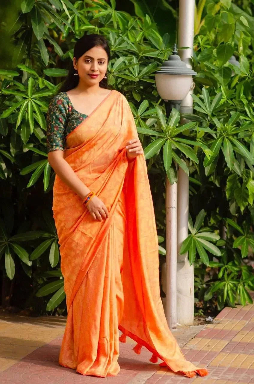 Model wearing orange soft silk jacquard saree with printed blouse standing outdoors near green foliage