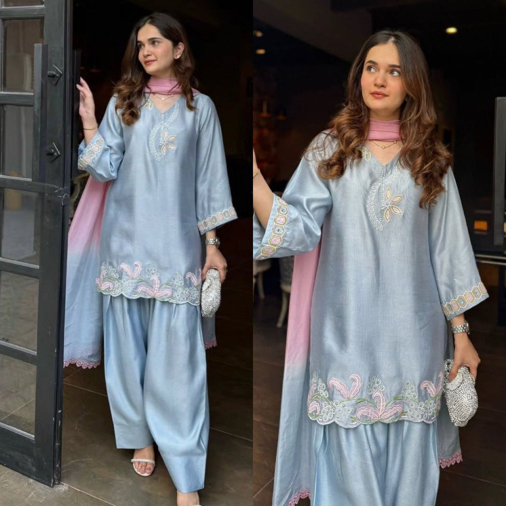 Woman posing in a light blue embroidered kurti and farshi salwar set with a pink ombre dupatta, holding a clutch, at an entrance.
