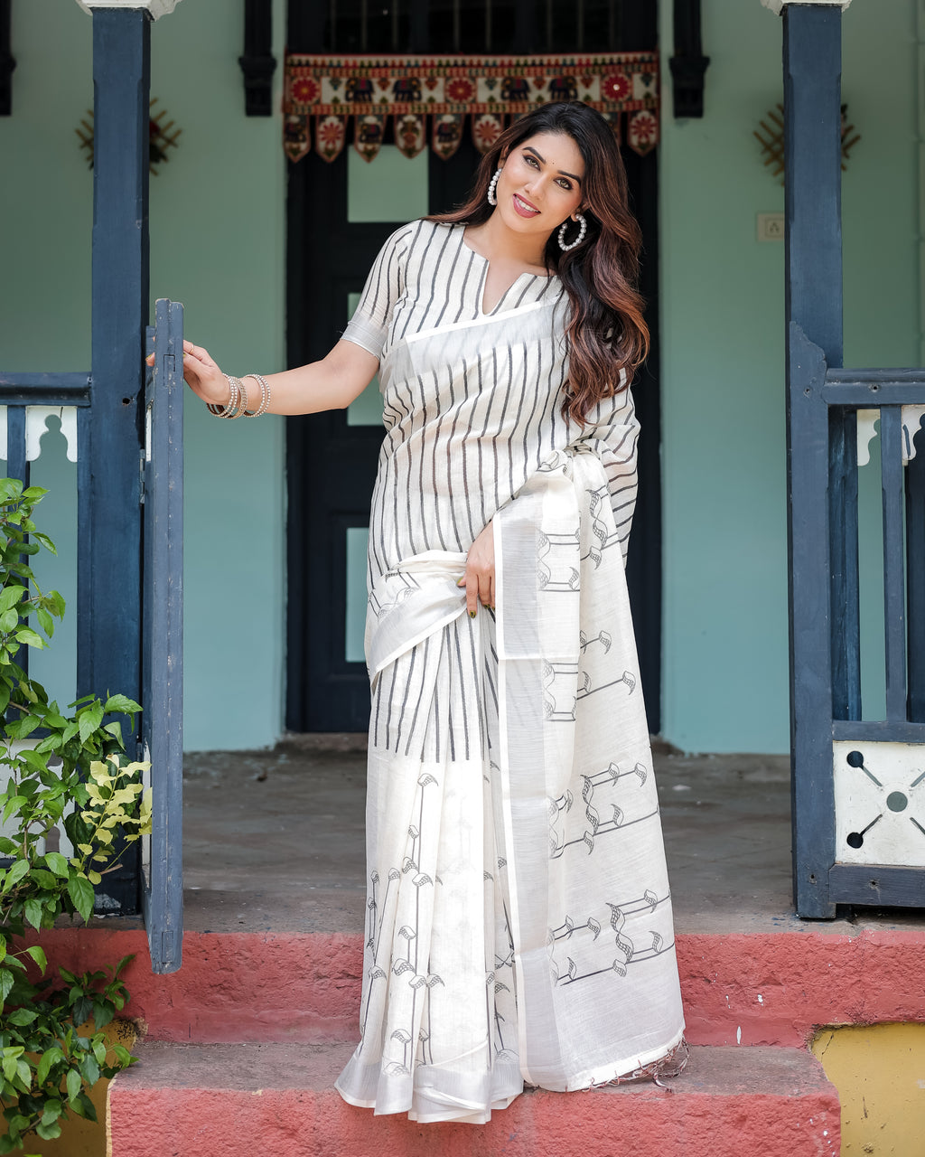 Woman posing in a white and black striped linen saree with tassels, front view, standing at a doorway.