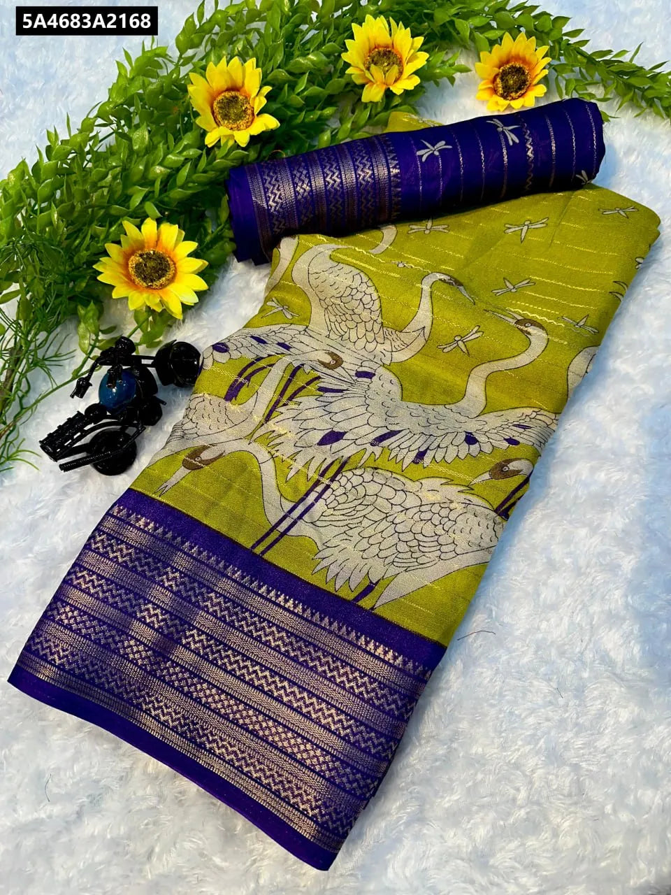 Flat lay of a vibrant lime green Mysore silk saree with intricate white crane weaving, a rich purple two-tone border, and a rolled matching purple blouse piece, set against a white background with decorative sunflowers.