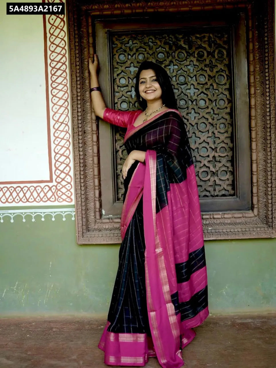 Front view of a smiling model wearing a Black Mysore Silk Saree with Pink border, standing by an ornate window.