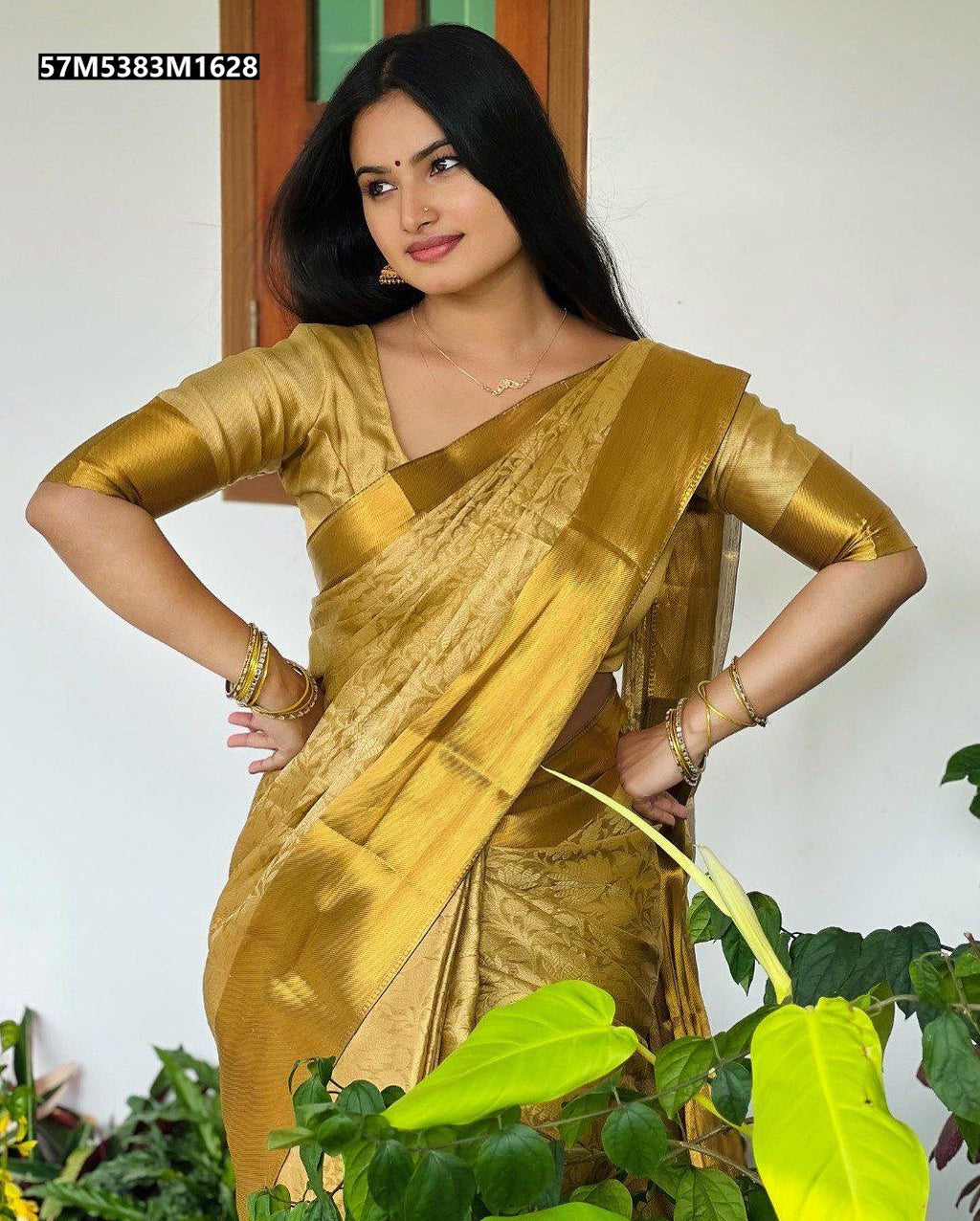 A beautiful woman with long black hair, wearing a shimmering golden tissue silk saree and matching blouse, striking a confident pose outdoors with hands on hips, smiling slightly, with green plants in the foreground.