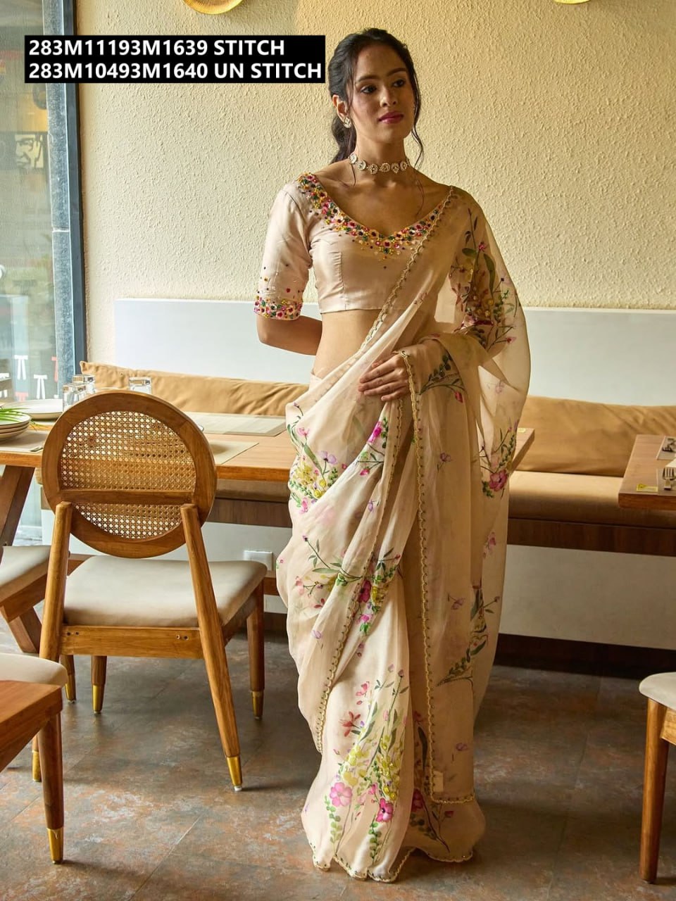 Woman modeling a peach Teby Silk floral printed saree with matching stitched raw silk blouse, featuring intricate colorful floral work, posing in a cafe.
