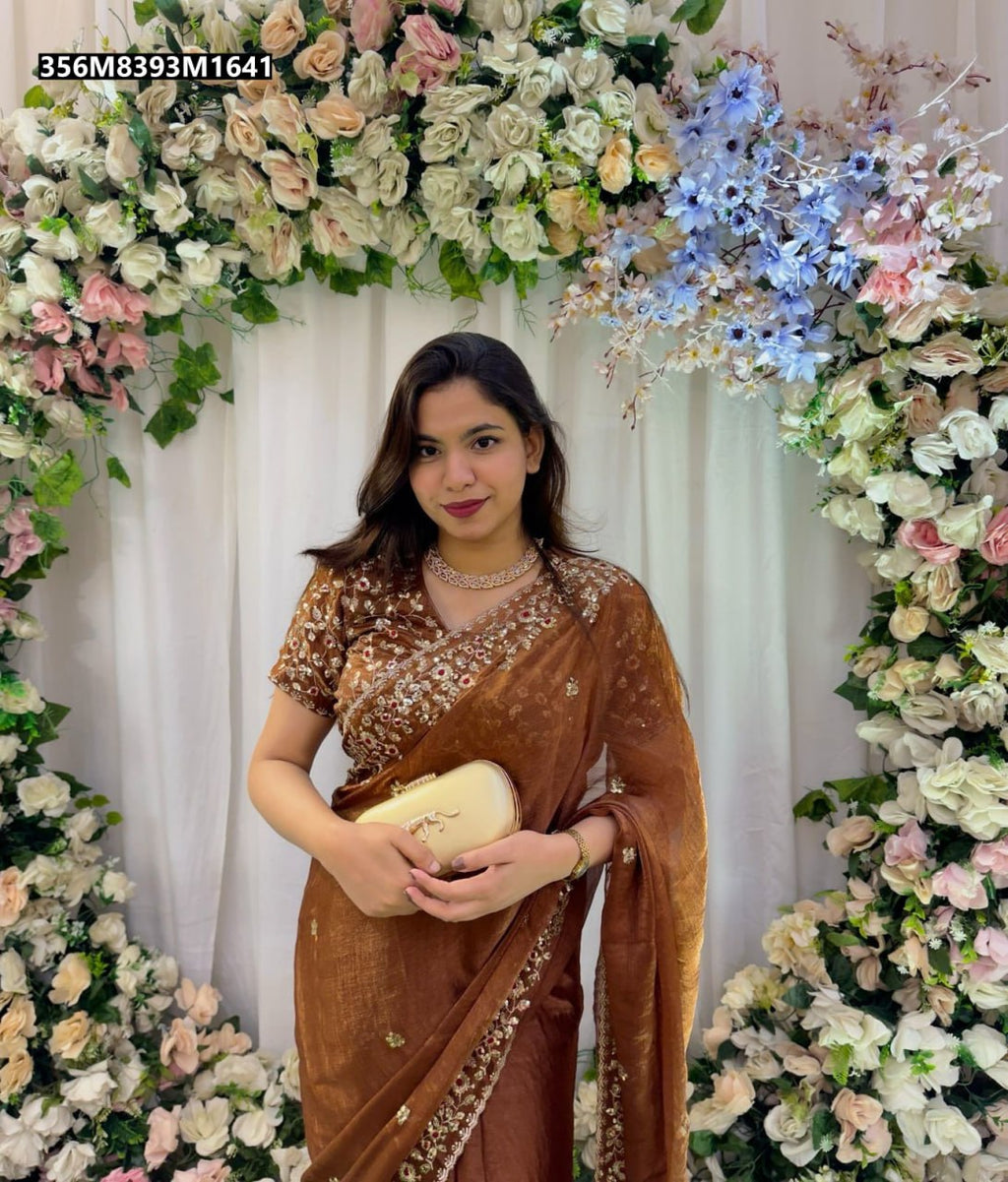 Model gracefully posing in a chocolate brown TRENDBUY sequin embroidered Frendy Chiffon saree with a matching stitched blouse, holding a gold clutch against a floral backdrop.