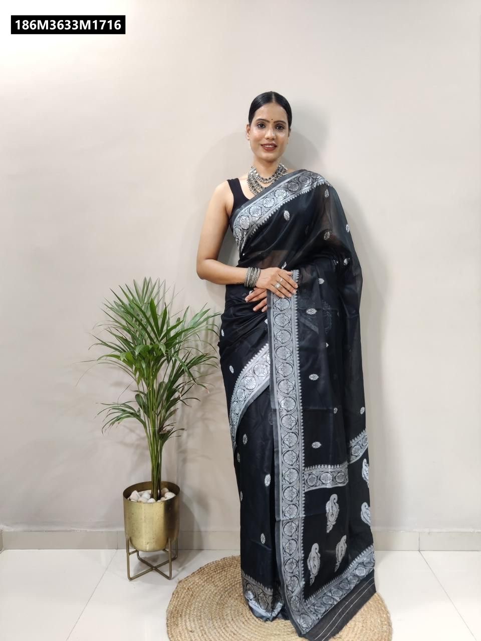 Full-length shot of a woman in a black soft lilan slab saree with intricate silver jacquard border, standing gracefully by a potted plant.