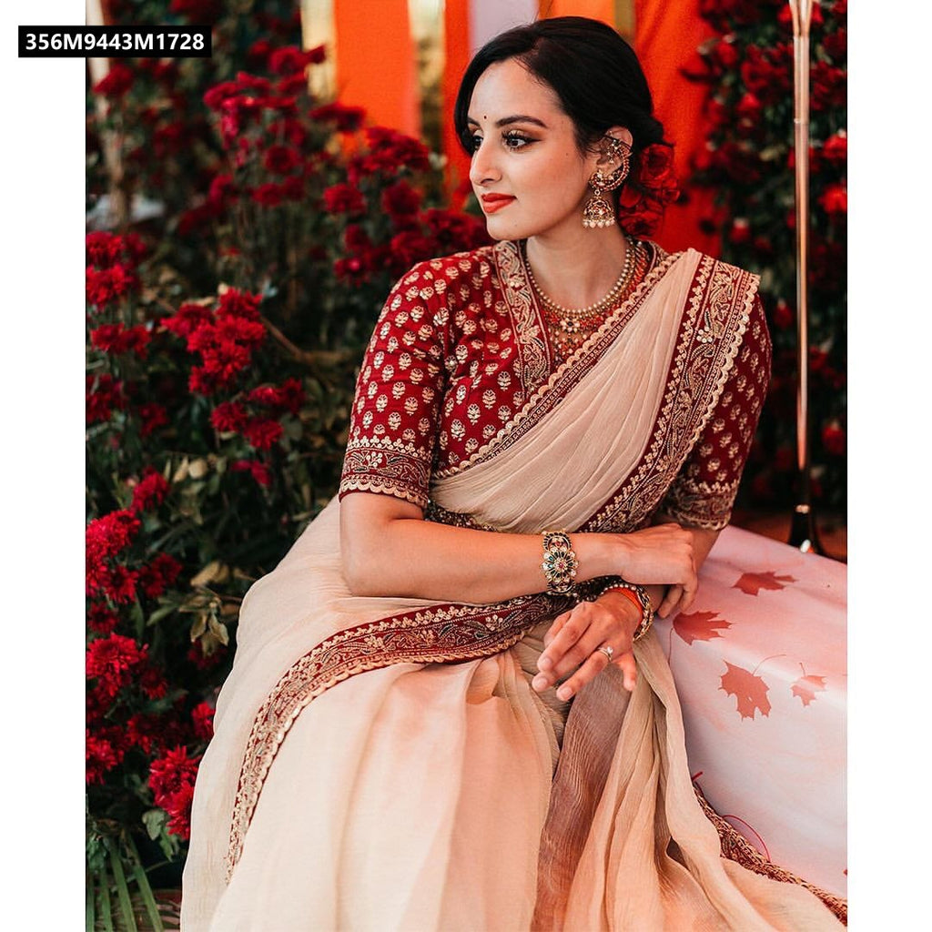 A beautiful woman seated elegantly, draped in the champagne gold saree with its rich red embroidered blouse. She is adorned with traditional jewelry and a red flower in her hair, looking thoughtfully to the side against a backdrop of vibrant red flowers.