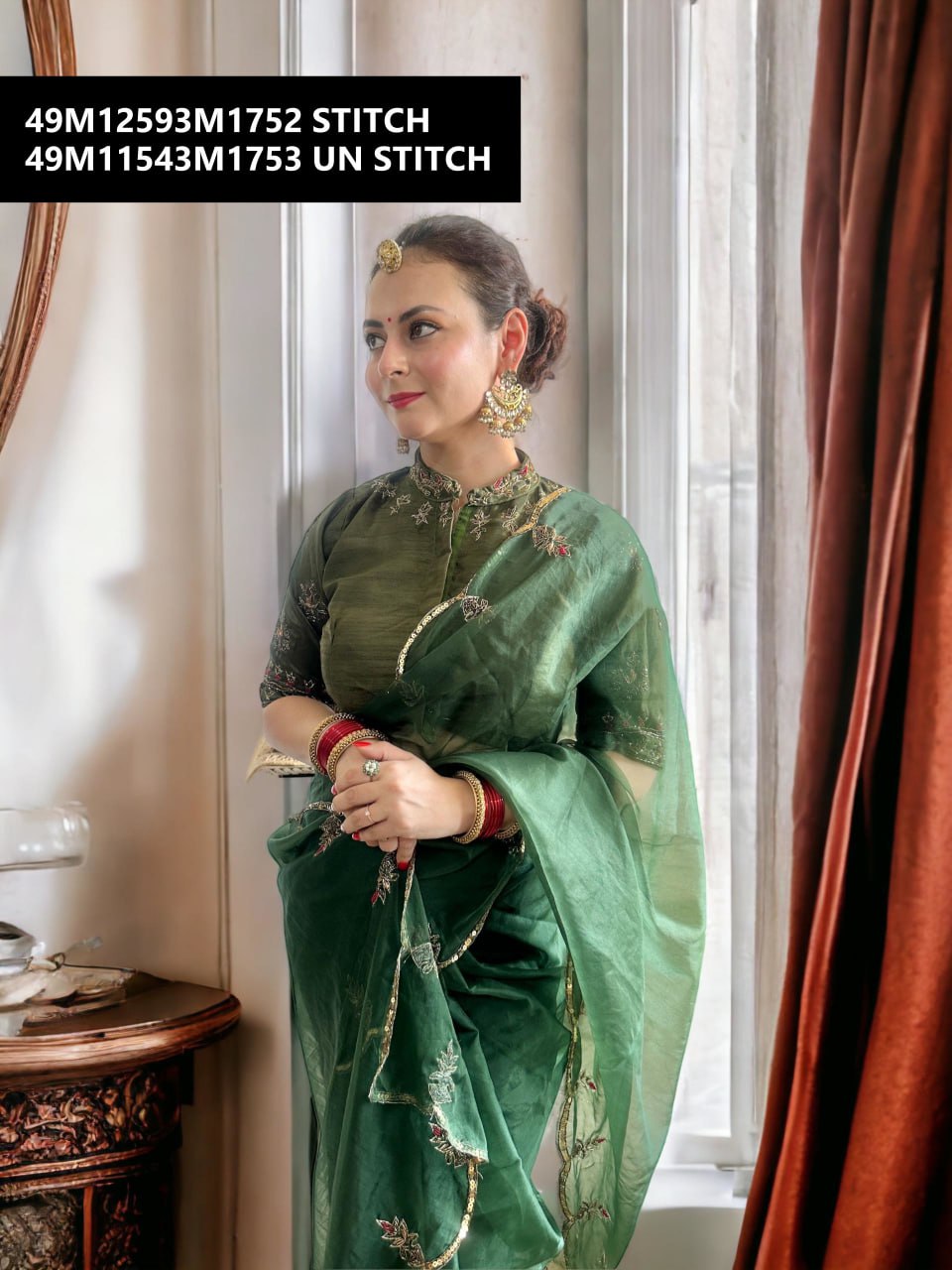 A woman elegantly draped in a TRENDBUY Emerald Green Organza Saree with intricate gold and red hand embroidery, posing by a window.