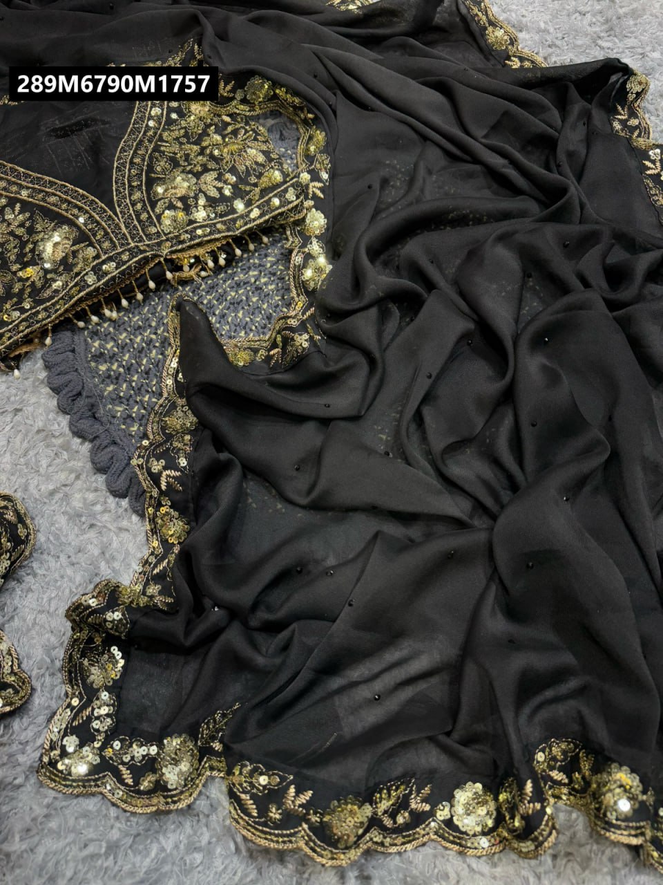 TRENDBUY black Rangoli silk saree with intricate gold sequin and thread embroidery on the border, laid flat on a grey textured surface.