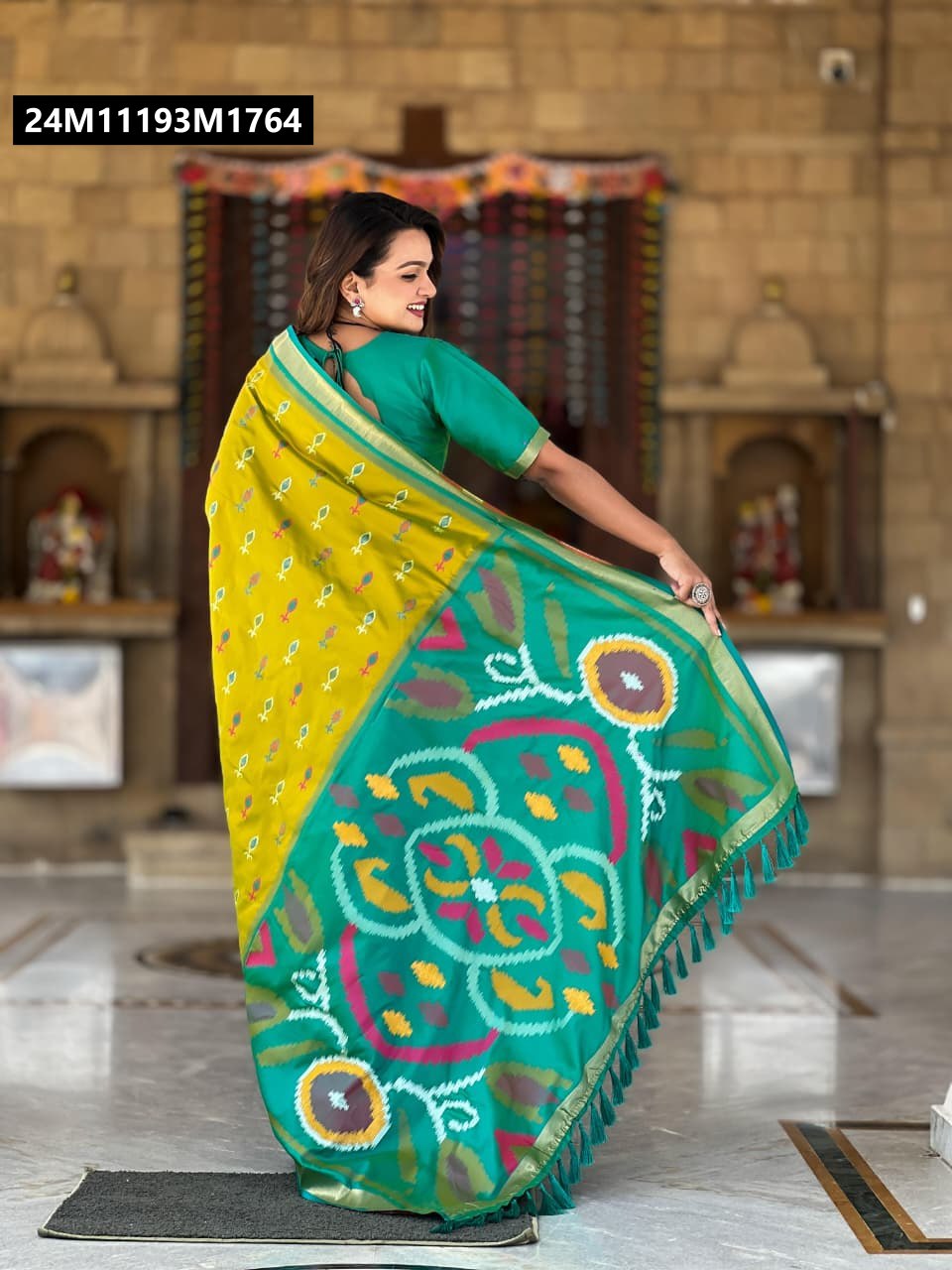Model smiling, posing elegantly with a Chartreuse and Teal soft silk saree featuring intricate Ikkat and Patola weaving, back view showing pallu with tassels.