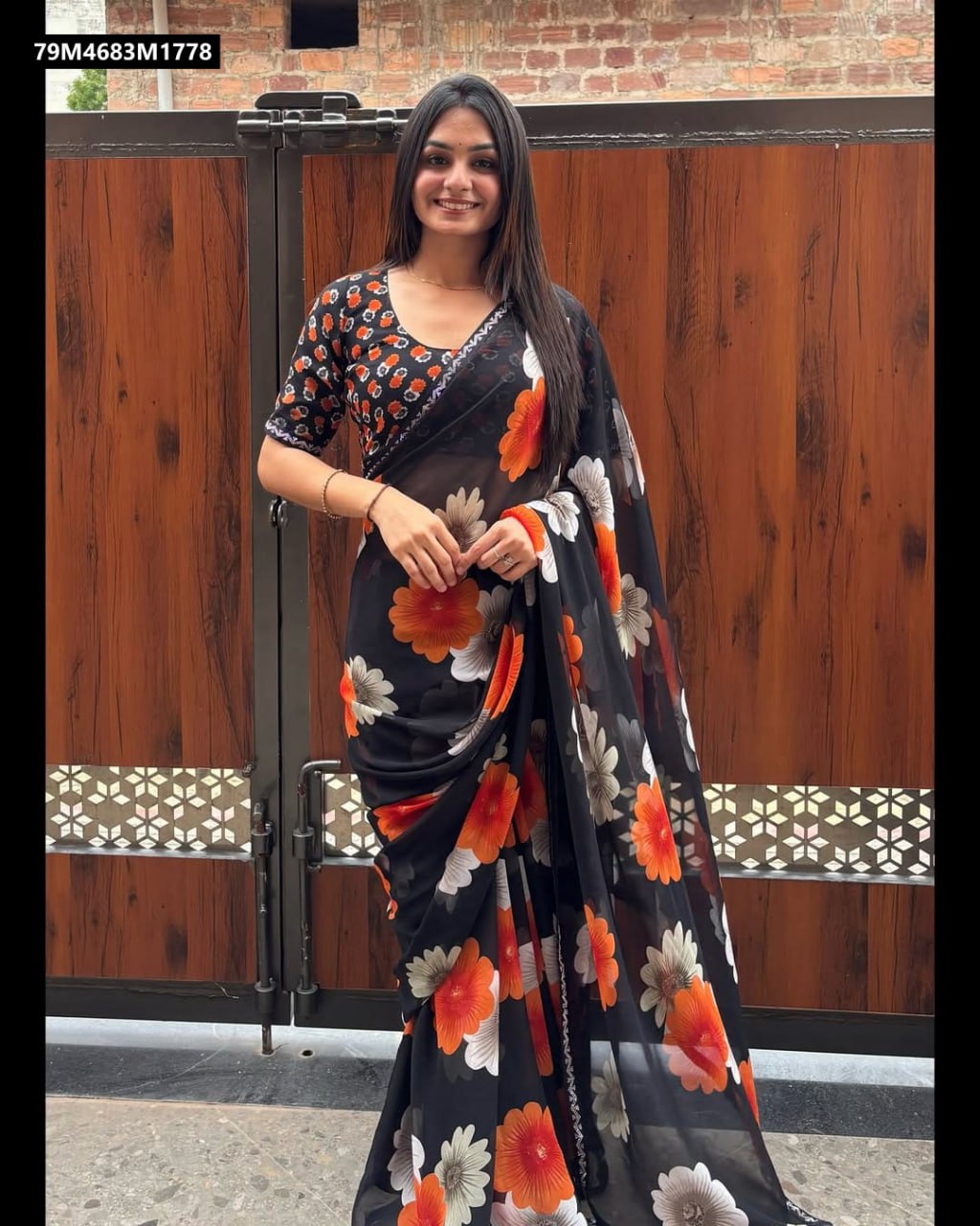 A smiling woman elegantly draped in a black Georgette saree with vibrant orange and white floral digital prints and a delicate Swarovski work border, posing in front of a rustic wooden door.