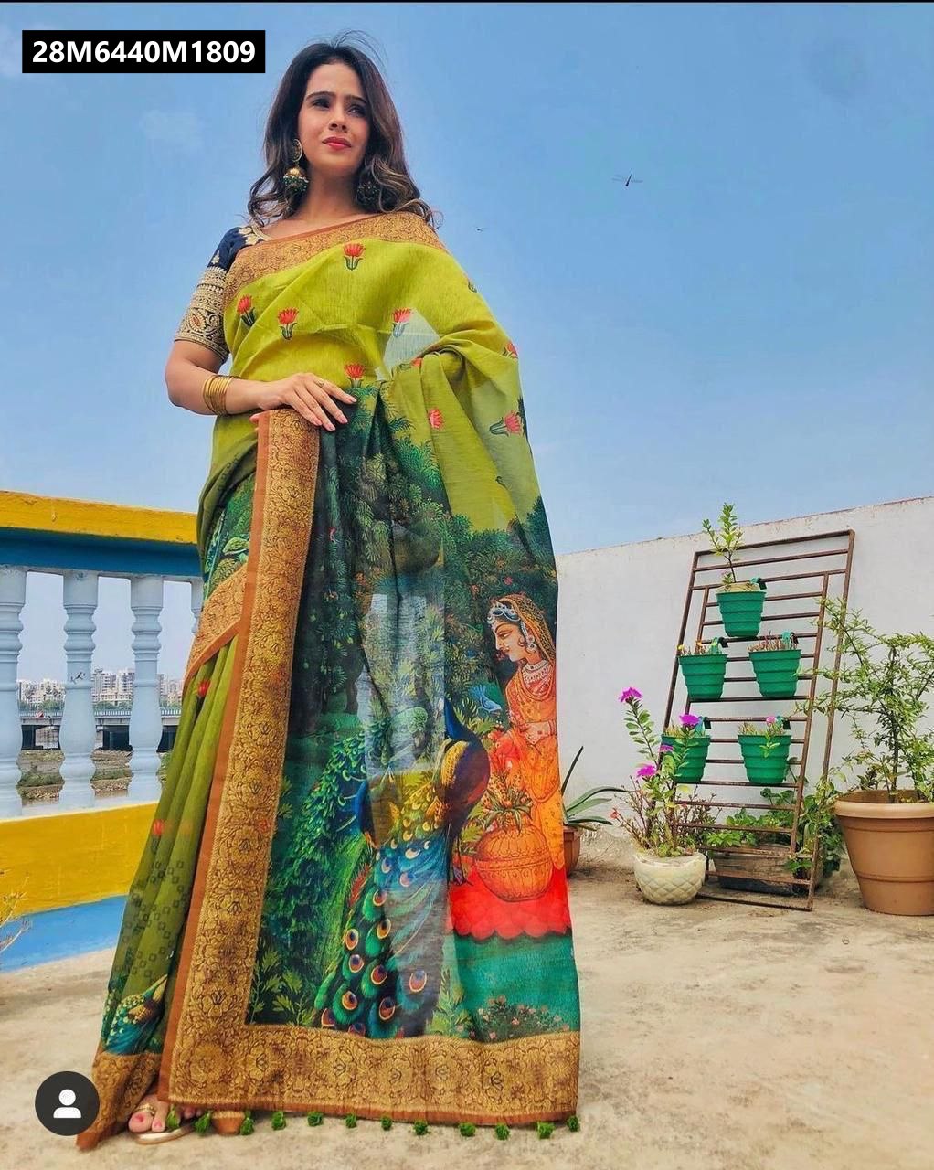 A model standing on a rooftop, elegantly draped in an olive green linen saree with a digital print depicting a woman and peacocks in a lush garden scene. The saree features a detailed Kalamkari-style border and green tassels.
