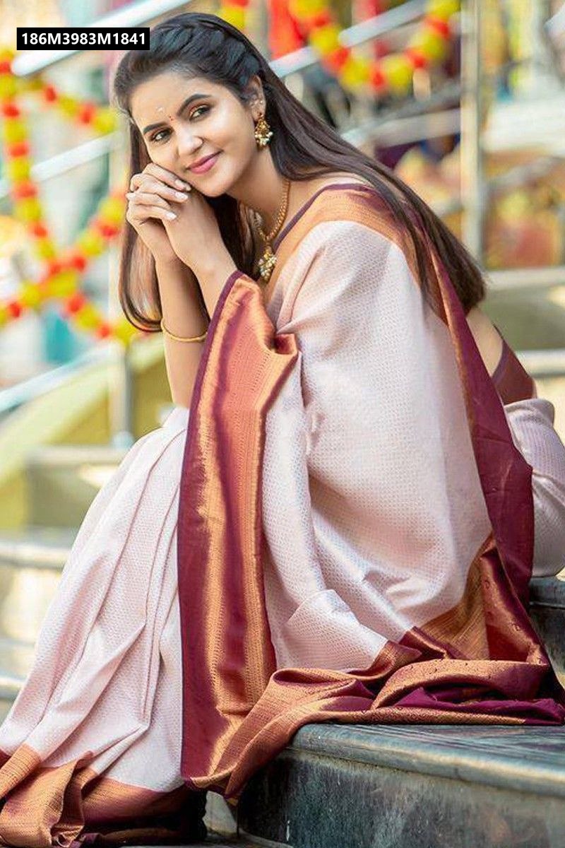 Smiling woman wearing a light beige Banarasi soft silk saree with a deep maroon zari-work border, sitting casually on steps.