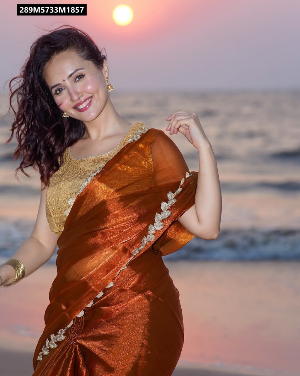 Close-up of a woman in a TRENDBUY coppery orange chiffon saree with a gold sequin blouse, smiling brightly with the sunset over the ocean.