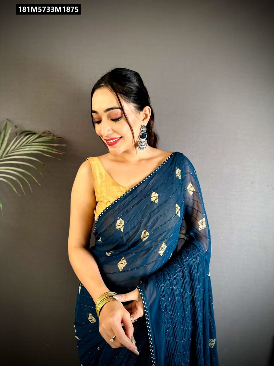 A smiling woman in a deep blue Fancy Georgette saree with gold foil diamond patterns and a gold blouse, adorned with traditional silver earrings and bangles, posing against a dark grey background.