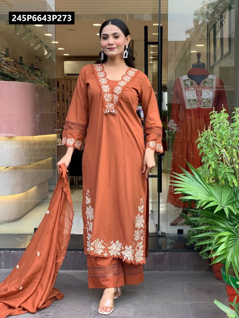 Full body shot of a woman modeling a rust-colored Roman fabric kurti set with white floral embroidery and cutwork lace, holding a matching dupatta.